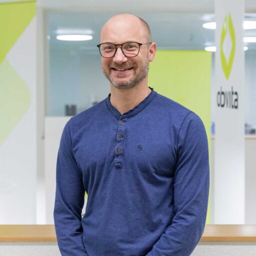 Ein Mann mit Brille und Bart lächelt und trägt ein blaues langärmeliges Henley-Shirt. Er steht in einem modernen Bürogebäude mit weißen und limettengrünen Akzenten und einem Logo mit der Aufschrift „dbvta“.