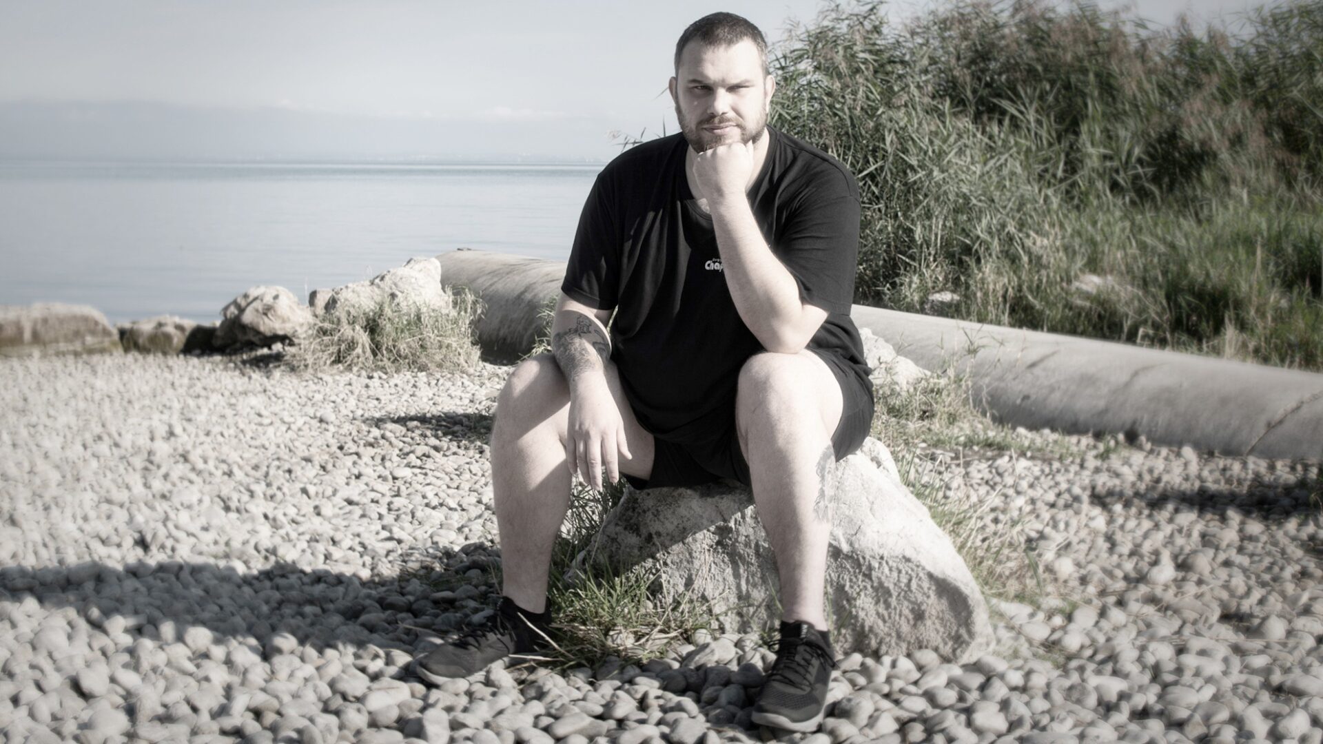Eine Person sitzt nachdenklich auf einem Felsen an einem felsigen Strand. Sie trägt ein schwarzes T-Shirt und Shorts und hat eine Hand am Kinn. Im Hintergrund sind hohe Gräser und ruhiges Wasser unter einem klaren Himmel zu sehen.