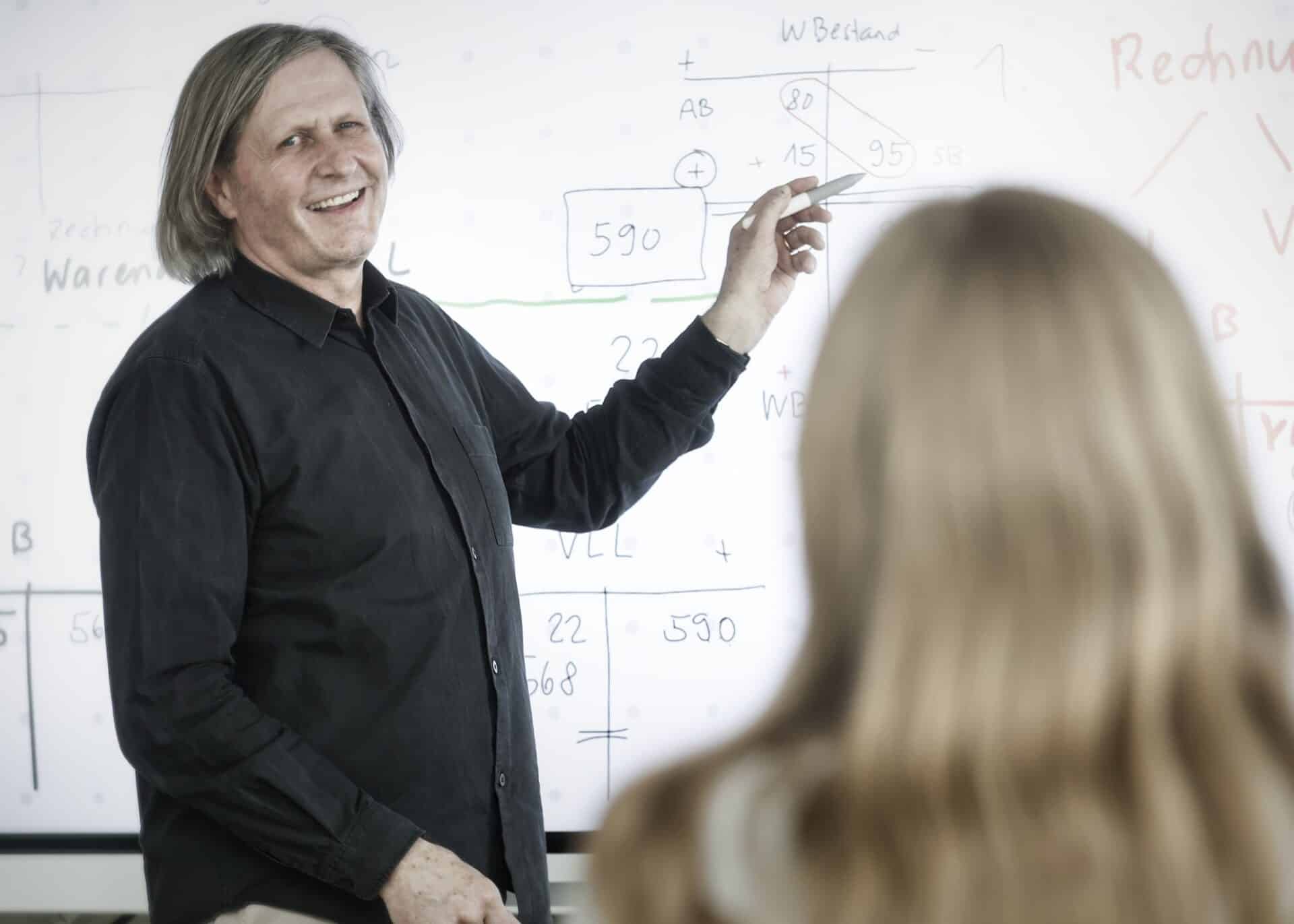 Eine Person mit langen Haaren und schwarzem Hemd steht lächelnd vor einer Tafel mit Diagrammen. Sie hält einen Marker in der Hand und zeigt auf die Tafel. Eine Person mit langen Haaren sitzt im Vordergrund und blickt auf die Tafel.