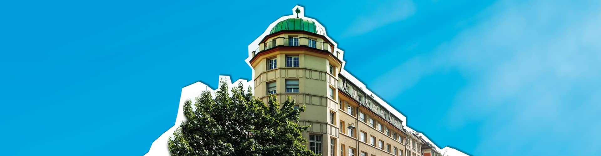 Ein hohes, historisches Gebäude mit einer grünen Kuppel an der abgerundeten Ecke steht unter einem klaren blauen Himmel. Ein belaubter Baum verdeckt teilweise den unteren Teil des Gebäudes. Das Gebäude hat helles Mauerwerk und zahlreiche Fenster.