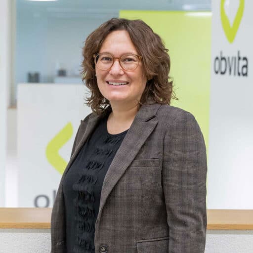 Eine Person mit schulterlangem lockigem Haar, Brille, braun kariertem Blazer und schwarzer Bluse steht lächelnd vor einem hellen Hintergrund, auf dem das Wort „obvita“ zu sehen ist.