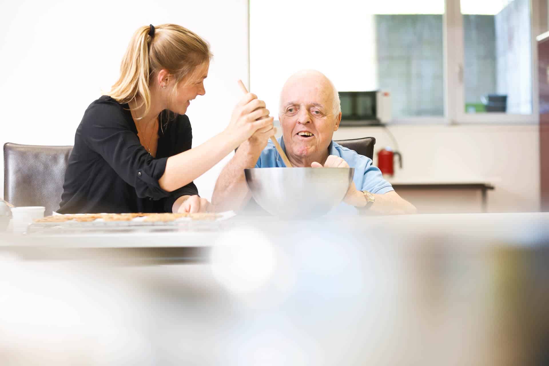 Eine junge Frau und ein älterer Mann in der obvita Talentwerkstatt backen gemeinsam. Sie rühren den Teig in einer grossen Schüssel und wirken konzentriert und fröhlich.
