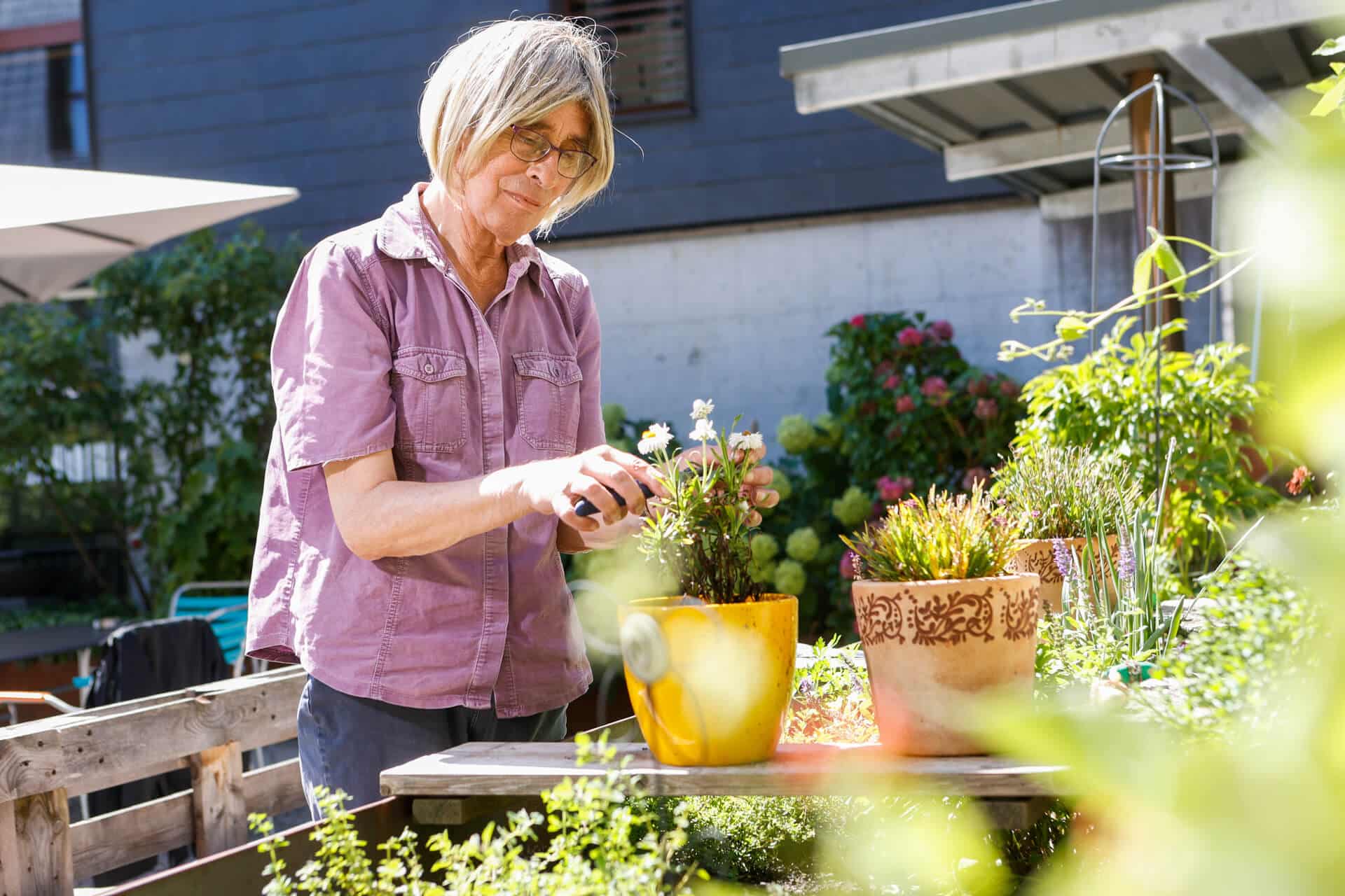 Eine Frau arbeitet in der obvita Talentwerkstatt im Garten und pflegt eine Pflanze in einem Blumentopf. Sie geniesst die Arbeit im Grünen, umgeben von üppiger Vegetation und farbenfrohen Blumentöpfen.