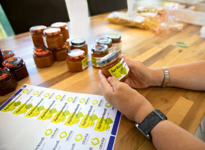 Eine Person in der obvita Talentwerkstatt etikettiert Gläser mit hausgemachter Aprikosenkonfitüre. Auf dem Tisch liegen weitere Konfitürengläser und Etiketten, was die Sorgfalt in der Produktverarbeitung unterstreicht.