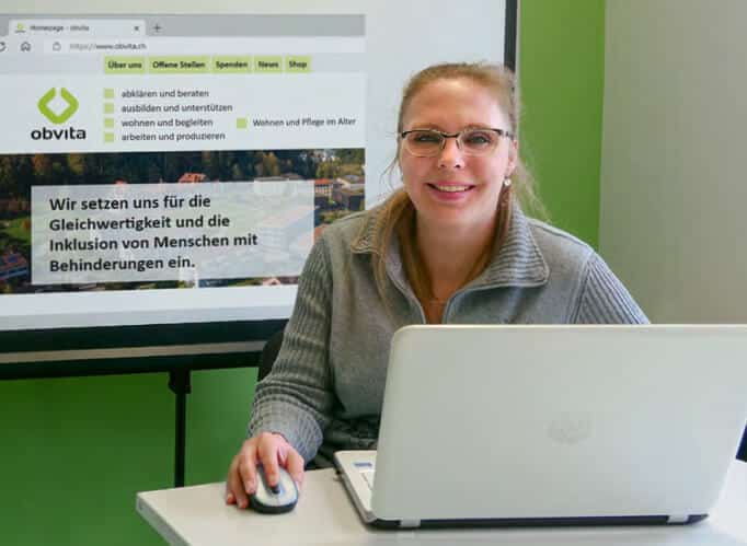Eine Mitarbeiterin in der obvita Talentwerkstatt sitzt lächelnd an einem Laptop. Im Hintergrund ist die Website von obvita auf einem Bildschirm zu sehen, mit einem Slogan zur Förderung der Inklusion von Menschen mit Behinderungen.