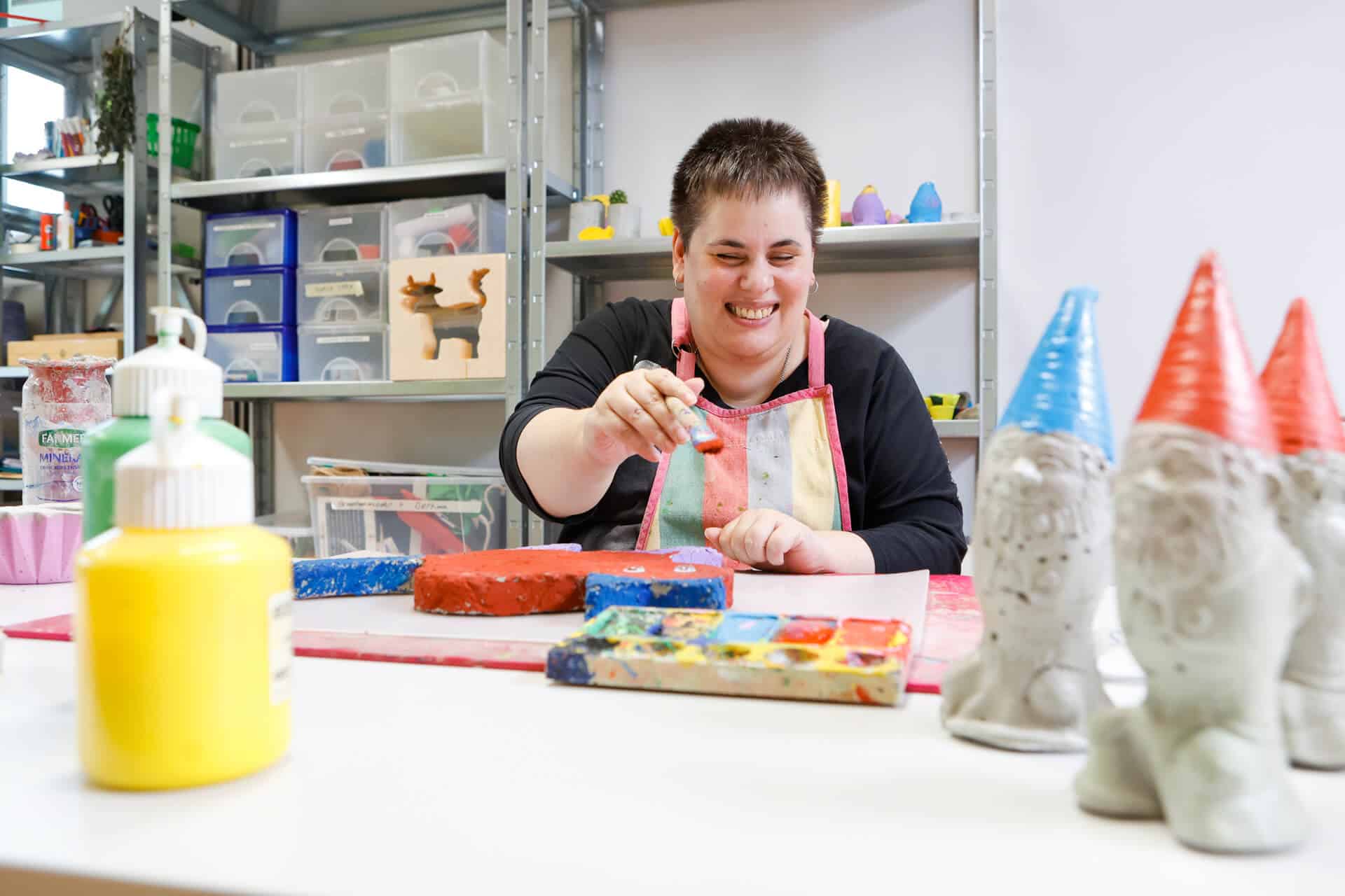 Eine Frau in der obvita Talentwerkstatt arbeitet kreativ mit Farben und Formen. Sie trägt eine Schürze und lächelt während des Gestaltungsprozesses. Im Vordergrund sind Farben und bemalte Figuren zu sehen, die die kreative Atmosphäre unterstreichen.