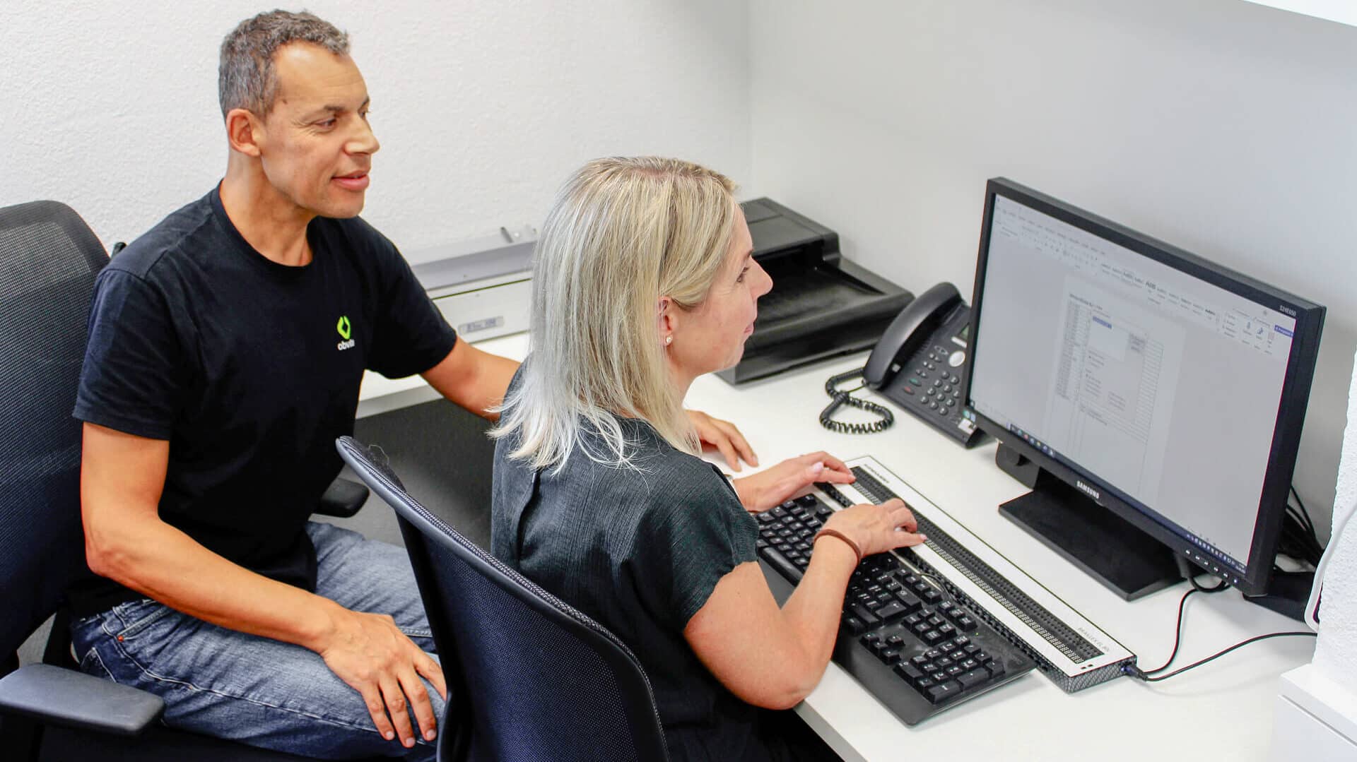 Ein Mann übt mit einer sehbeeinträchtigten Frau die Bedienung eines Computers.