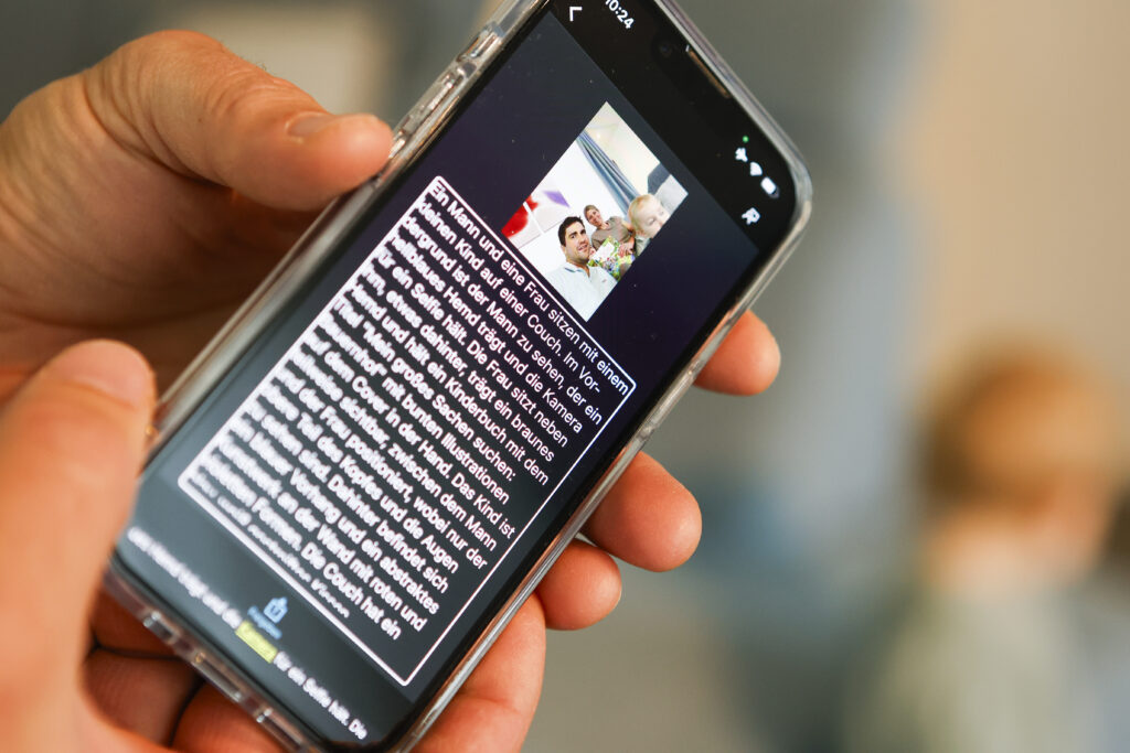 Eine Hand hält ein Smartphone, auf dem ein Familienfoto und eine lange Nachricht in deutscher Sprache in weißer Schrift auf schwarzem Hintergrund zu sehen sind, mit einem verschwommenen Kind im Hintergrund.