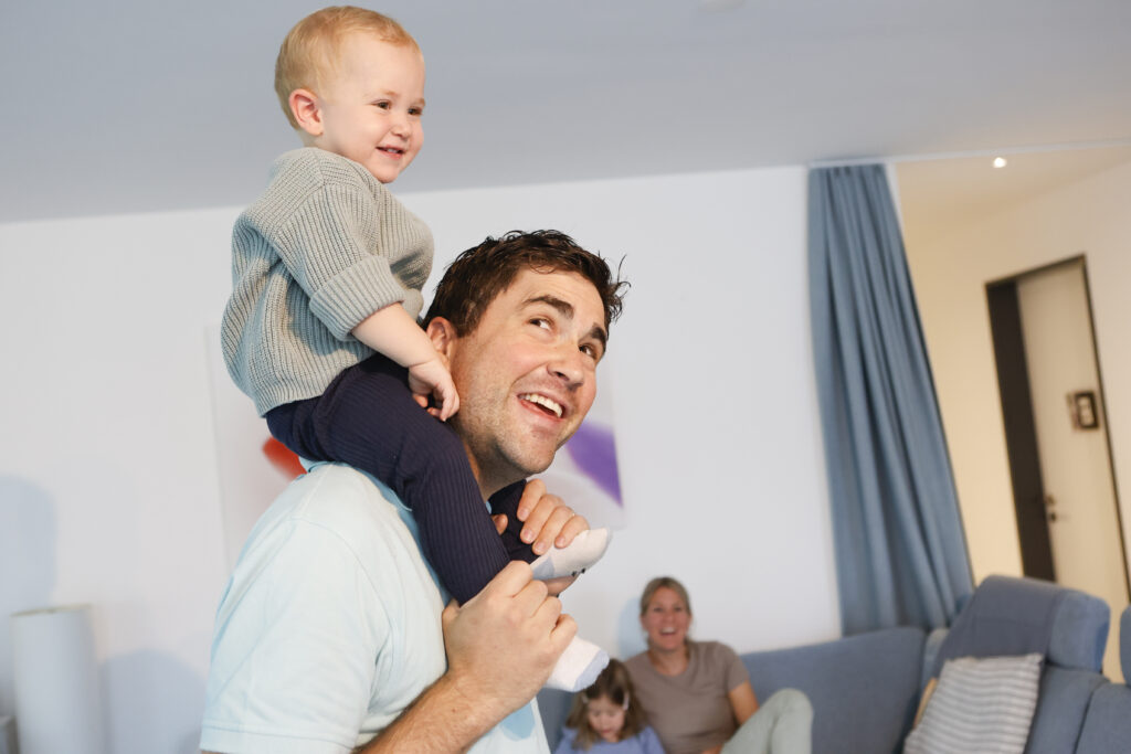Ein lächelnder Mann trägt ein glückliches Kleinkind auf seinen Schultern in einem Haus. Im Hintergrund sitzen eine Frau und ein weiteres Kind auf einem Sofa, alle wirken entspannt und fröhlich in einem hellen Wohnzimmer.