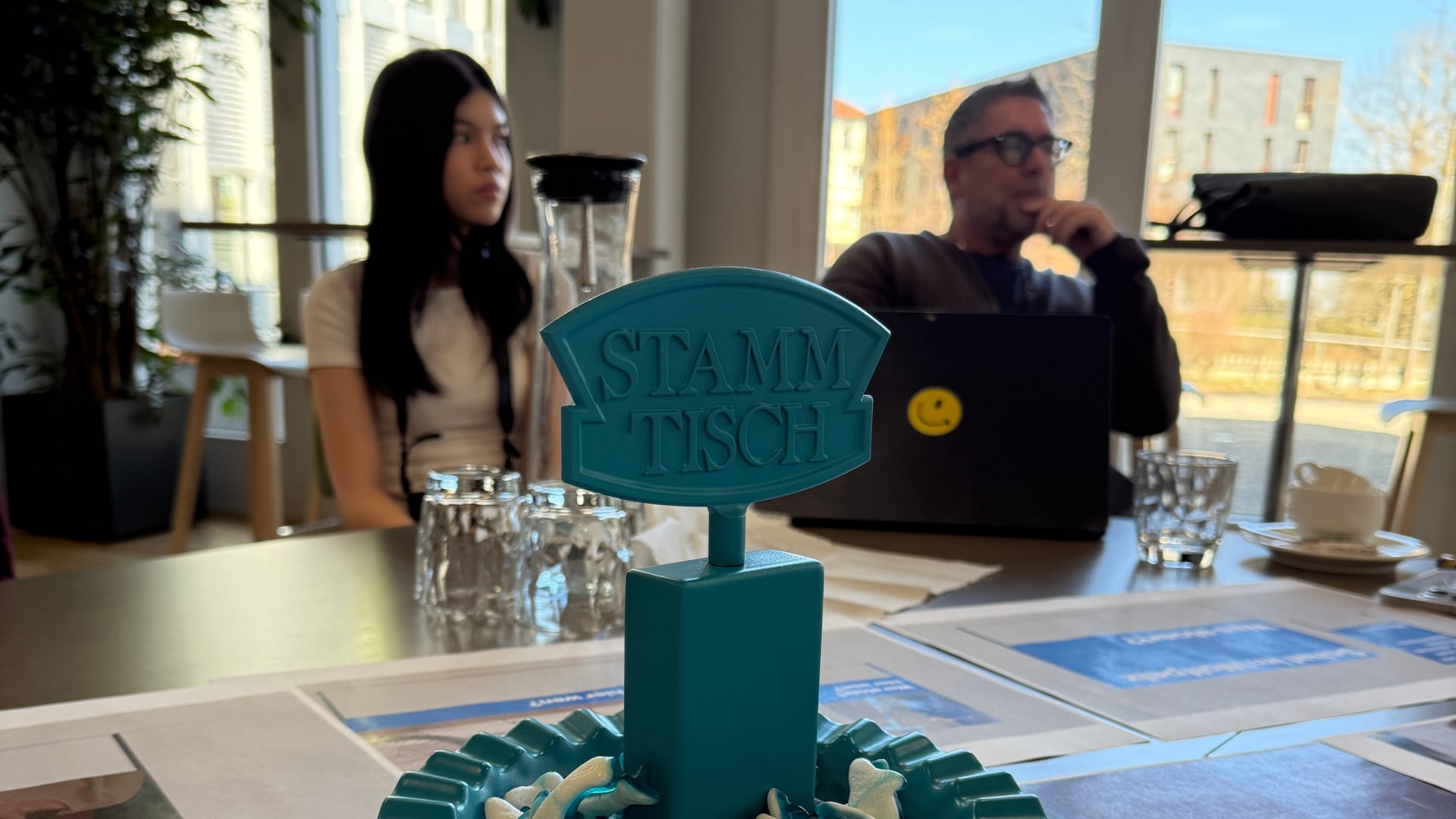 Eine Nahaufnahme eines blauen „Stammtisch“-Schildes auf einem Tisch, umgeben von Papieren und Gläsern. Im Hintergrund sitzen zwei Personen, eine Frau und ein Mann mit einem Laptop, auf dem ein Smiley-Aufkleber angebracht ist. Grosse Fenster geben den Blick auf Gebäude und Bäume frei.