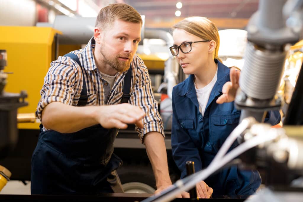 Zwei Ingenieure in Arbeitsuniformen besprechen und untersuchen Maschinen in einer Industrieanlage. Der Mann deutet auf die Anlage, während die Frau aufmerksam zuhört. Beide konzentrieren sich auf die technischen Details.