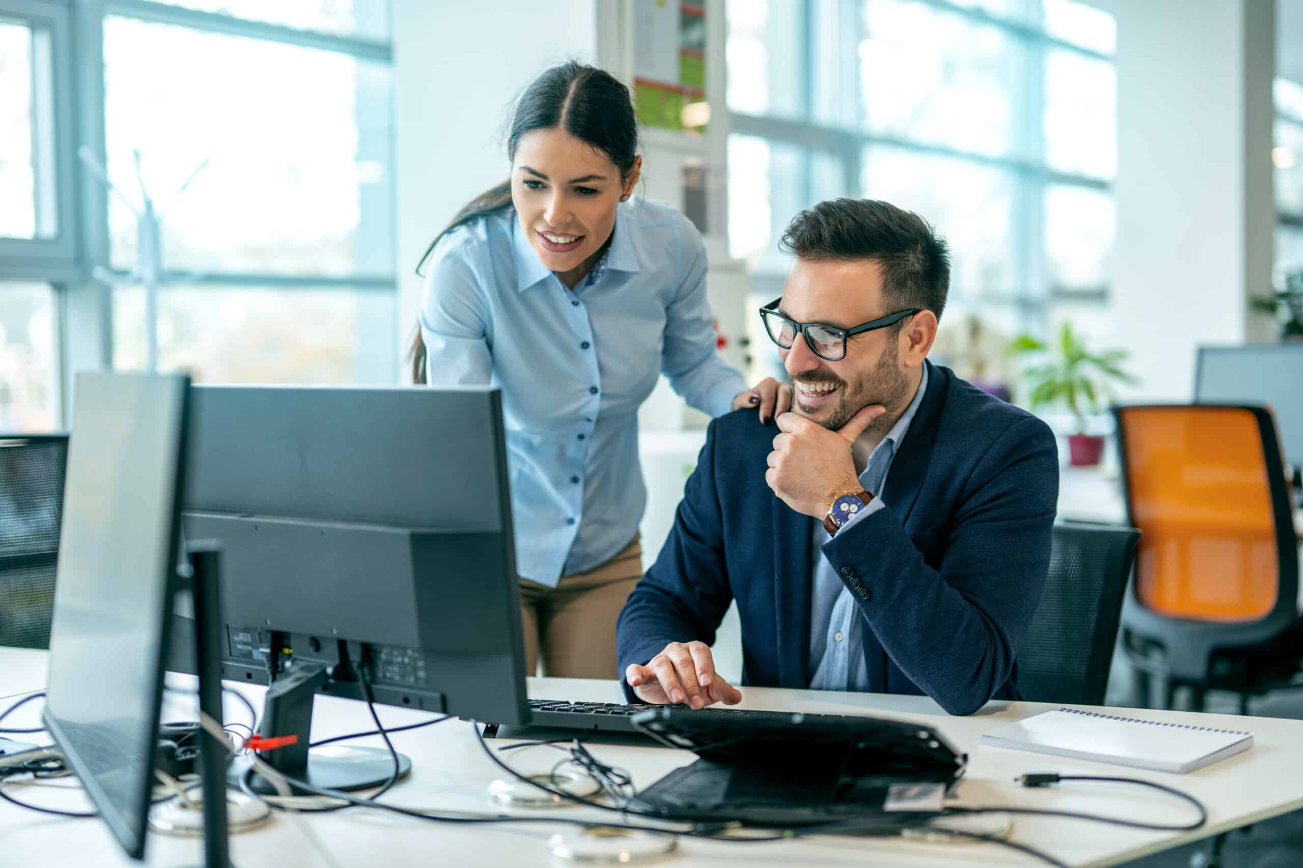 Zwei Kollegen in einem Büro. Einer sitzt lächelnd an einem Schreibtisch, benutzt einen Computer, während der andere daneben steht und auf den Bildschirm schaut. Beide wirken engagiert und kooperativ.