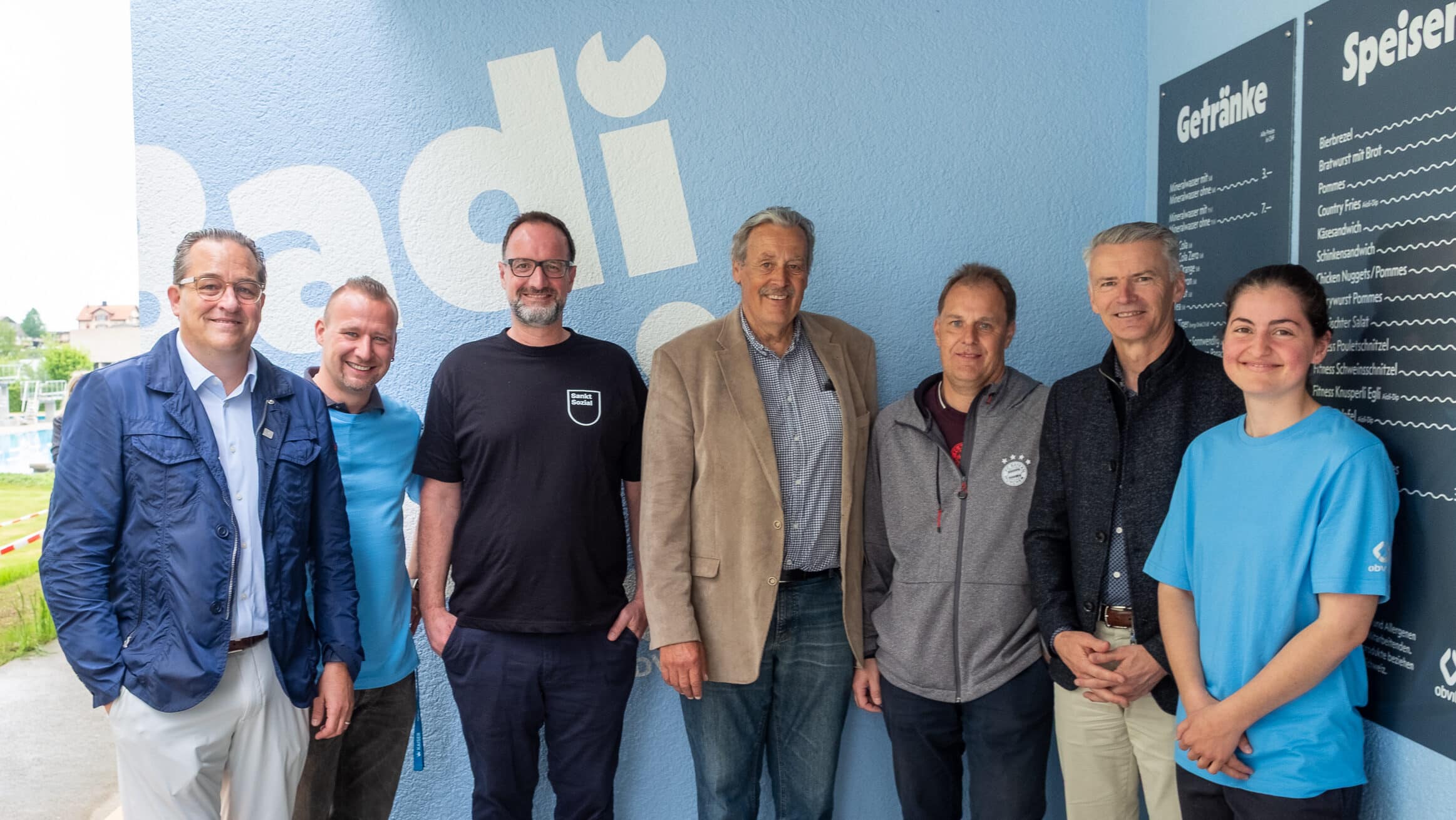 Sieben Menschen stehen lächelnd zusammen in einem Raum neben einer hellblauen Wand. Dahinter hängt eine Speisekarte und in großen Buchstaben steht „Badi“. Sie sind leger gekleidet und durch ein Fenster ist draussen eine Rasenfläche zu sehen. Im Bild: v.l.n.r: Patrik Läser, Präsident obvita Ostschweizerischer Blindenfürsorgeverein, Sven Hillmann, Koordinationsperson Zentralküche obvita bruggwald51, Christoph Tobler, Co-Geschäftsleiter obvita, Bruno Bovelli, Präsident der Verwaltung Badi Sonnenrein Wittenbach, Daniel Worni, Bereichsleiter Bau und Infrastruktur Gemeinde Wittenbach, Peter Bruhin, Gemeindepräsident Wittenbach, Nicole Geiger, Leiterin BadiKafi Wittenbach.