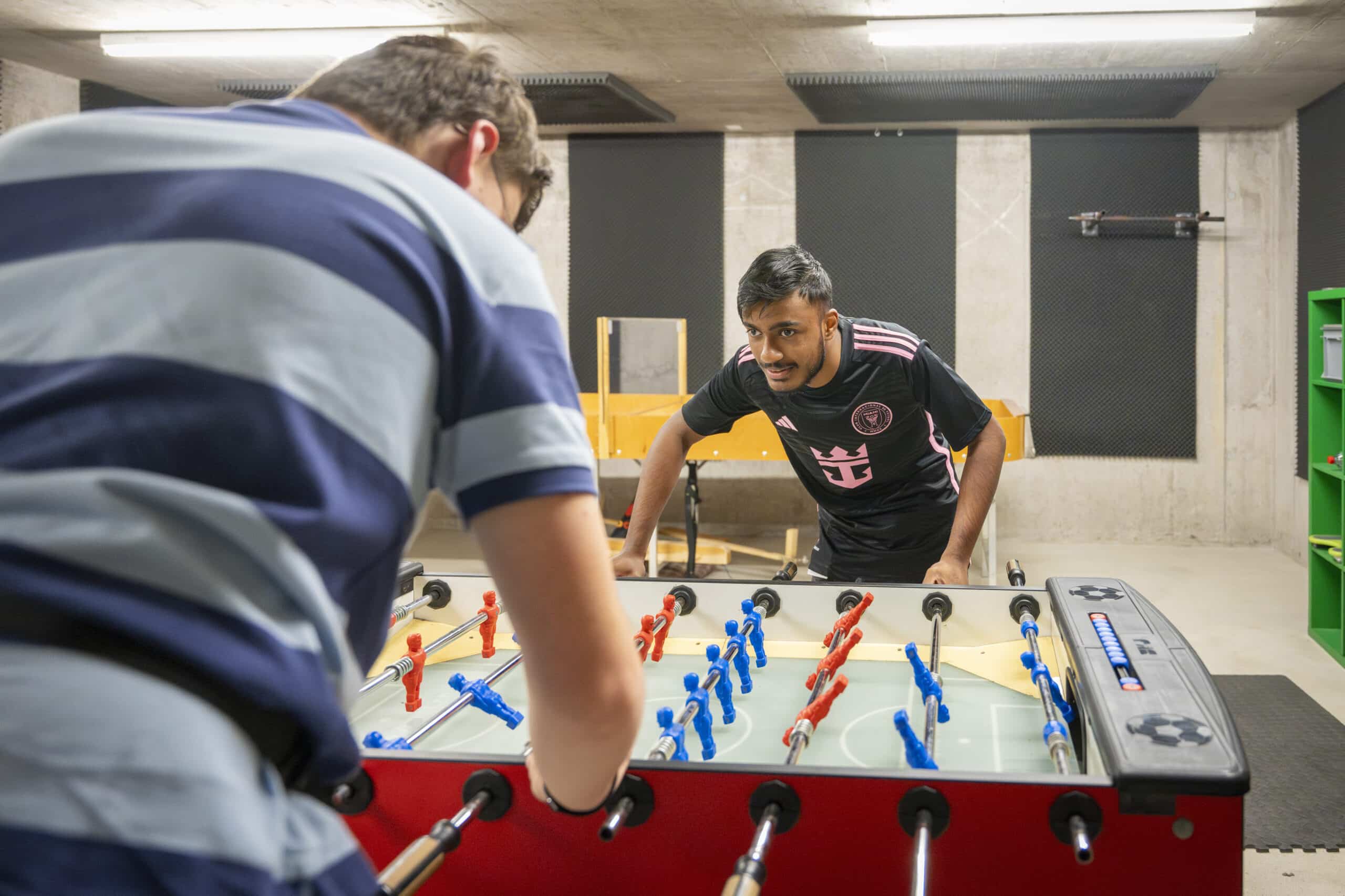 Zwei Männer spielen konzentriert Tischfußball in einem hell erleuchteten Raum mit Betonwänden. Einer trägt ein gestreiftes Hemd, der andere ein schwarzes Sporttrikot. Der bunte Tischfußballtisch ist der Mittelpunkt der Szene.