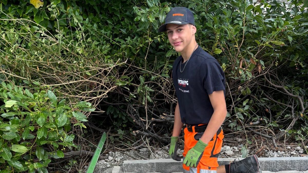 Eine Person in orangefarbener Sicherheitskleidung, mit Handschuhen und einer Mütze kniet auf neu verlegten Pflastersteinen und hält eine Fliese. Sie arbeitet neben grünen Sträuchern, in der Nähe befinden sich einige Pflasterwerkzeuge und Materialien.