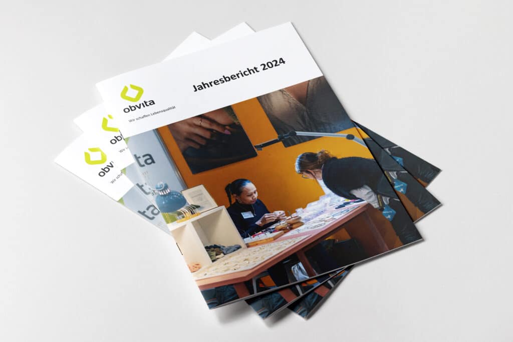 Ein Stapel Jahresberichtshefte mit dem Titel „Jahresbericht 2024“ von obvita, mit einem Titelbild von zwei Personen, die in einer warmen, kreativen Umgebung an einem Tisch mit Bastelmaterialien arbeiten.