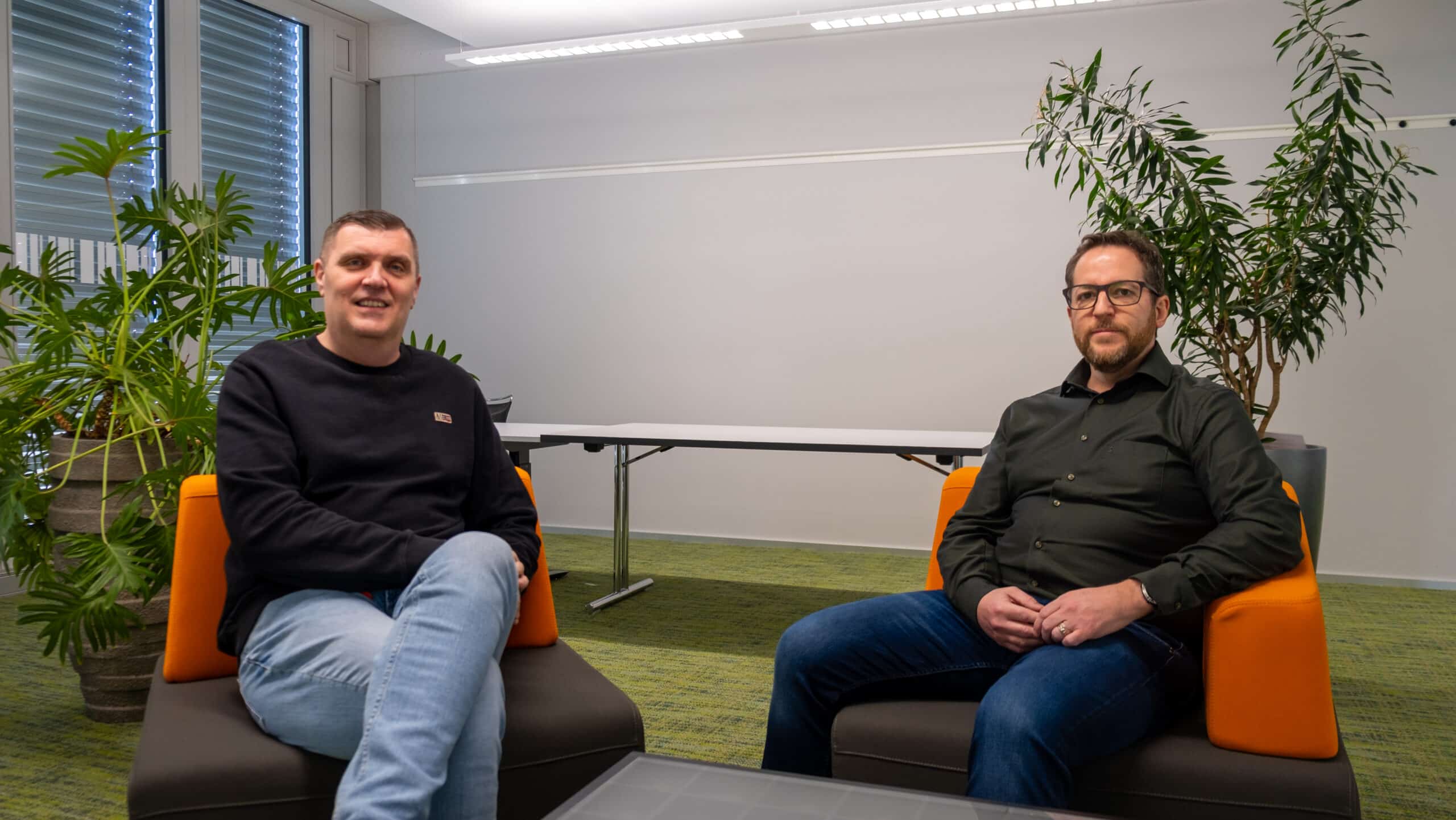 Zwei Männer sitzen auf orangefarbenen Stühlen in einem modernen Büro mit grünem Teppich, Pflanzen und einem Tisch im Hintergrund. Beide sind leger gekleidet, schauen in die Kamera und haben einen neutralen Gesichtsausdruck.