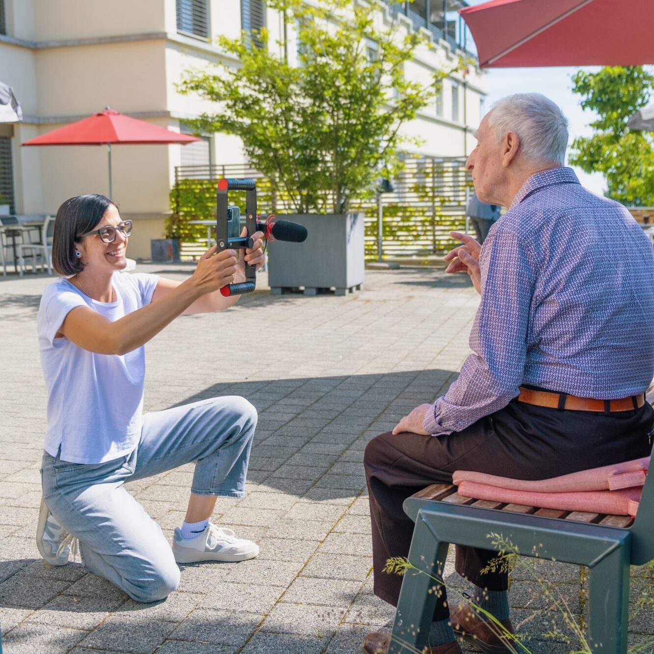 Eine Frau kniet im Freien und interviewt mit einem Smartphone und einem Mikrofon einen älteren Mann, der auf einer Bank sitzt. Sie befinden sich in einem sonnigen Innenhof mit modernen Gebäuden, Pflanzen und roten Regenschirmen im Hintergrund.