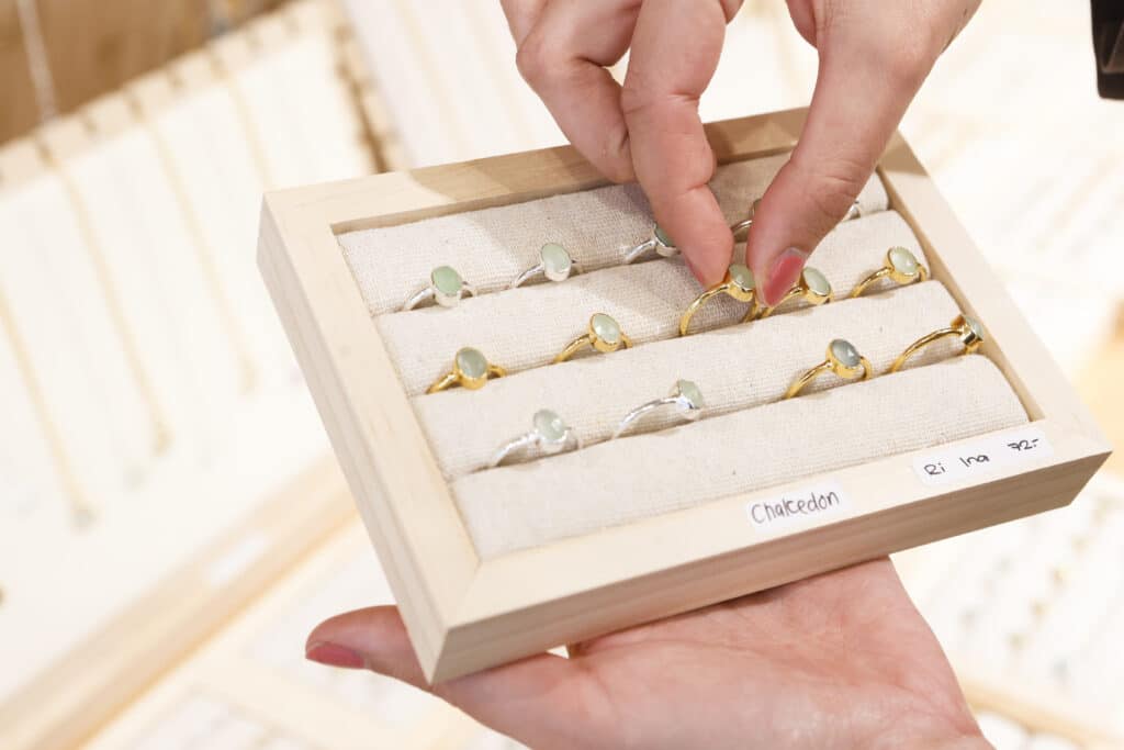 Eine Hand hält ein Tablett mit mehreren Goldringen, die jeweils mit einem hellgrünen Edelstein besetzt sind, während eine andere Hand auf einen Ring zeigt. Auf dem Tablett steht „Chalcedon“.