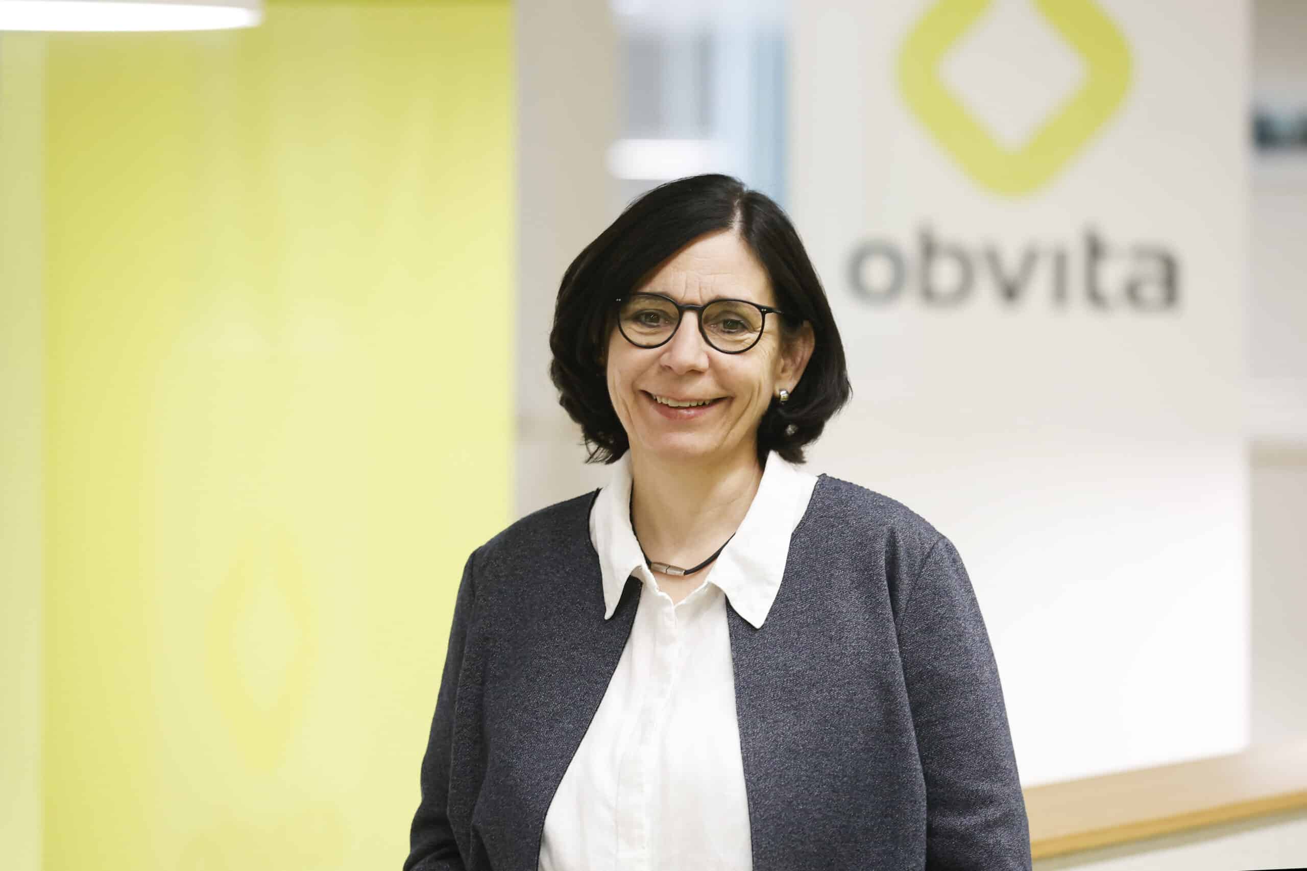 Eine Frau mit kurzen dunklen Haaren und Brille, die eine weiße Bluse und einen grauen Blazer trägt, steht lächelnd vor einem verschwommenen Hintergrund mit dem "obvita"-Logo.
