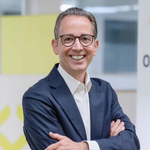 Ein lächelnder Mann mit Brille, der einen dunklen Blazer und ein weißes Hemd trägt, steht mit verschränkten Armen in einem hellen, modernen Büro.