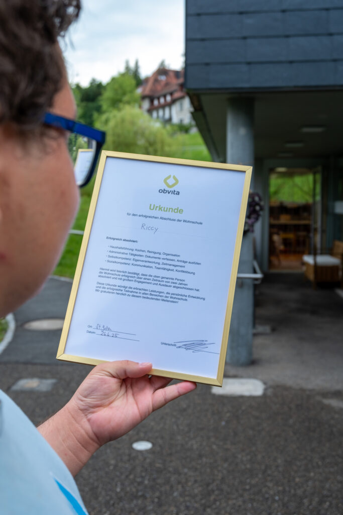 Eine Person mit kurzen lockigen Haaren und Brille hält eine gerahmte Urkunde vor einem modernen Gebäude, während im Hintergrund Grünflächen und ein traditionelles Haus zu sehen sind. Die Urkunde trägt den Titel "Urkunde.