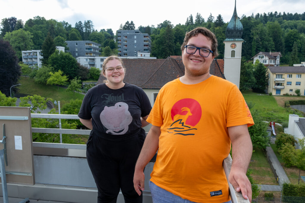 Zwei Personen stehen lächelnd auf einem Dach mit einem malerischen Hintergrund aus Bäumen, Häusern und einer Kirche mit einem Uhrenturm. Die Person auf der linken Seite trägt ein schwarzes Hemd, die Person auf der rechten Seite ein orangefarbenes Hemd mit einem Berg- und Sonnenmotiv.