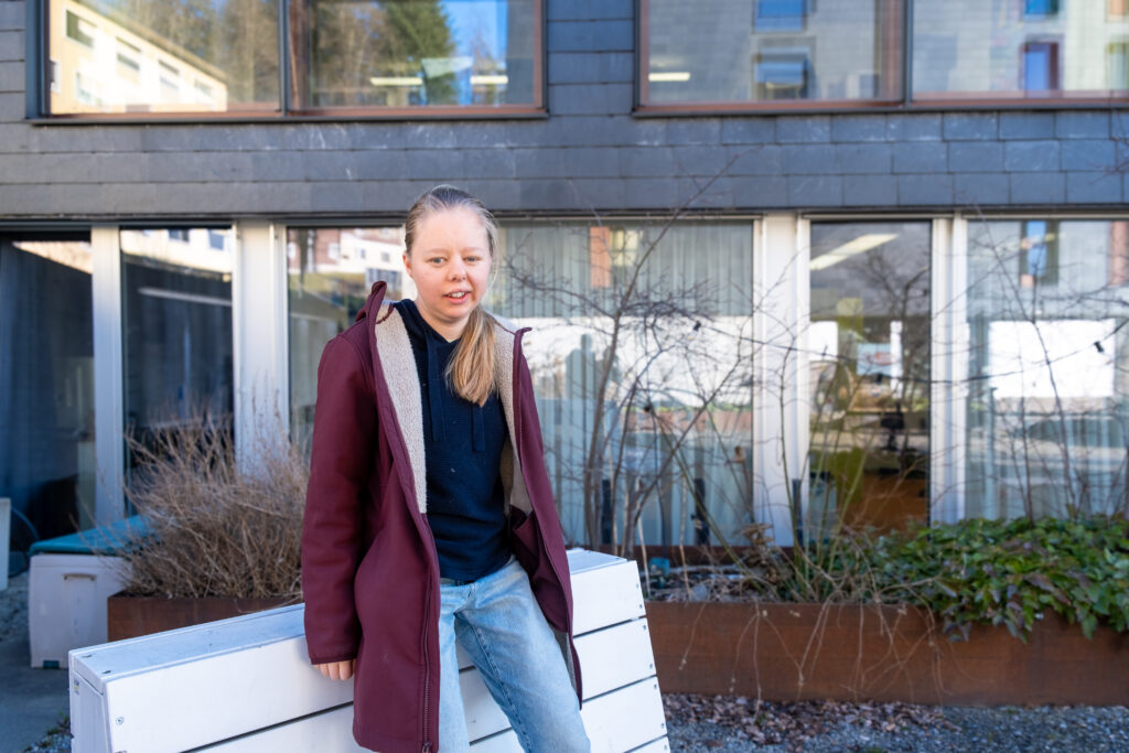 Eine junge Person mit langen blonden Haaren, einem kastanienbraunen Mantel und blauen Jeans sitzt auf einer weißen Bank im Freien vor einem modernen Gebäude mit großen Fenstern und Pflanzen.