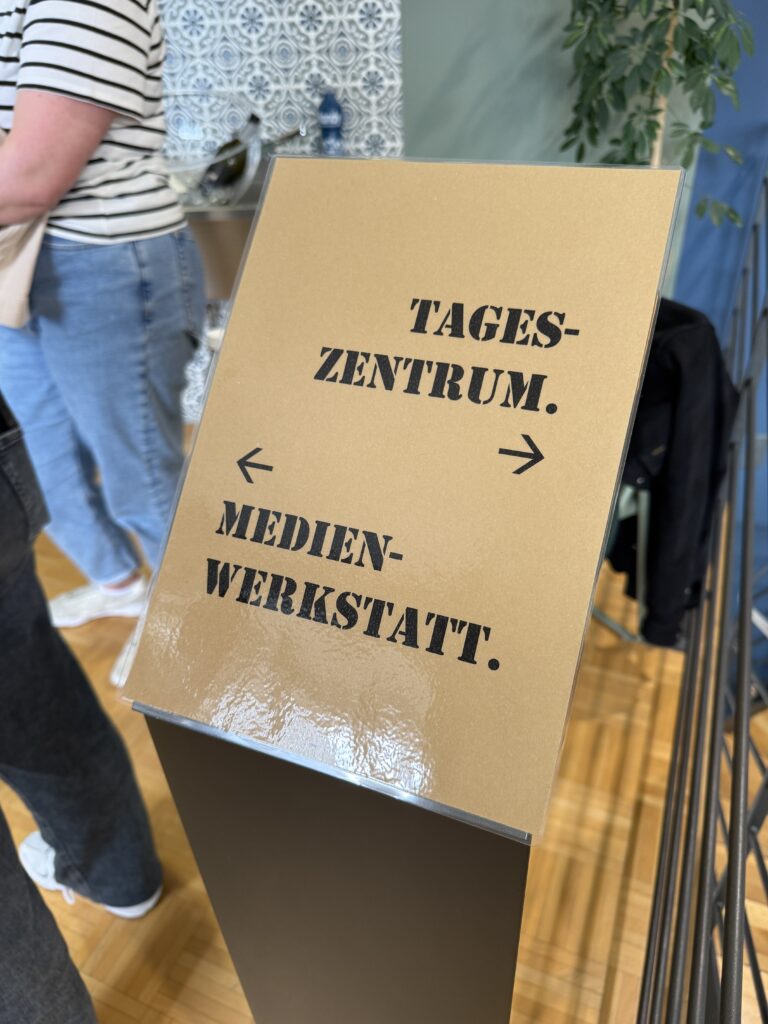 Ein Schild mit schwarzer Schablonenschrift in deutscher Sprache weist links auf "Tageszentrum" und rechts auf "Medienwerkstatt" hin. Neben dem Schild ist ein Teil einer Person in einem gestreiften Hemd und einer Jeans zu sehen.