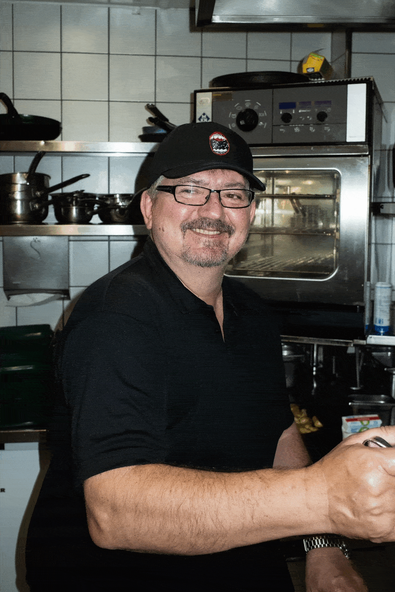 Ein Mann mit Brille, schwarzem Polohemd und schwarzer Mütze lächelt, während er in einer Großküche steht und ein Küchengerät hält. Im Hintergrund sind Töpfe und ein Ofen zu sehen.
