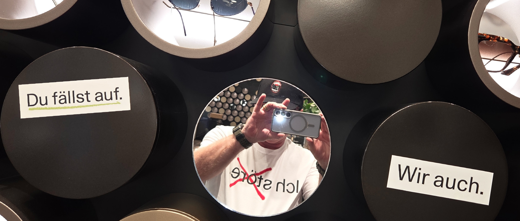 Eine Person macht ein Spiegel-Selfie, umgeben von runden Sonnenbrillen-Displays. Auf Deutsch lauten die Aufkleber "Du fällst auf."  "Ich störe" (durchgestrichen), und "Wir auch."