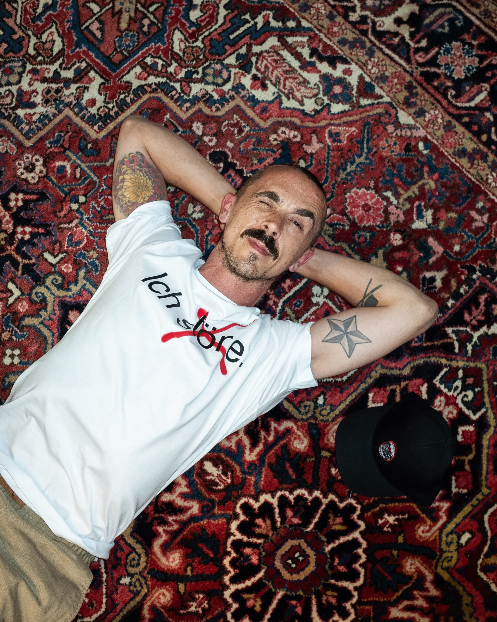Ein Mann mit Tätowierungen liegt auf einem gemusterten Teppich, die Arme hinter dem Kopf verschränkt, trägt ein weißes T-Shirt mit der Aufschrift "love", auf dem "love" durchgestrichen ist, und eine schwarze Mütze liegt neben ihm.