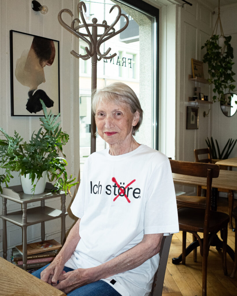 Eine ältere Frau mit kurzen grauen Haaren sitzt an einem Tisch in einem gemütlichen Café. Sie trägt ein weißes T-Shirt mit der Aufschrift "Ich störe" und einem roten X über dem Wort. Sie ist umgeben von Pflanzen, Büchern und Wandkunst.