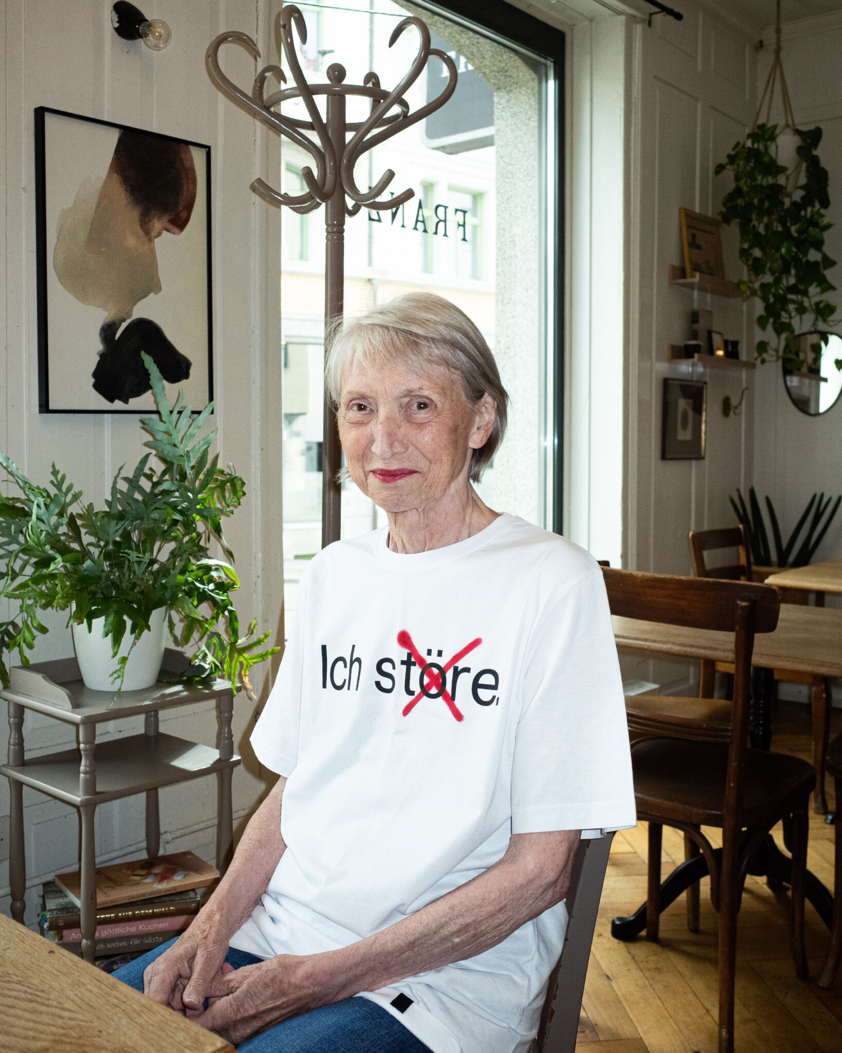 Eine ältere Frau mit kurzen grauen Haaren sitzt an einem Tisch in einem gemütlichen Café. Sie trägt ein weißes T-Shirt mit der Aufschrift "Ich störe" und einem roten X über dem Wort. Sie ist umgeben von Pflanzen, Büchern und Wandkunst.