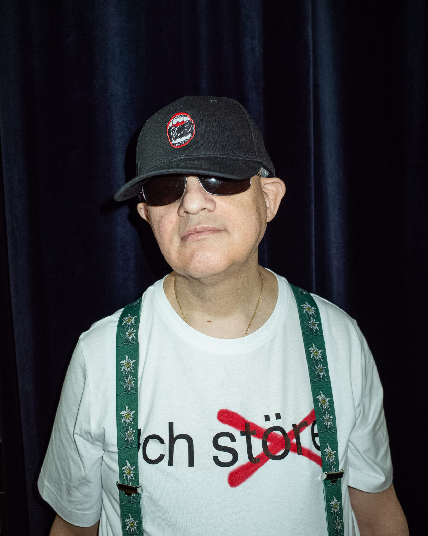 Ein Mann, der eine schwarze Baseballkappe, eine dunkle Sonnenbrille und ein weißes T-Shirt mit der Aufschrift "ich störe" trägt, steht vor einem dunklen Vorhang. Er trägt außerdem grüne Hosenträger mit einem Blumenmuster.