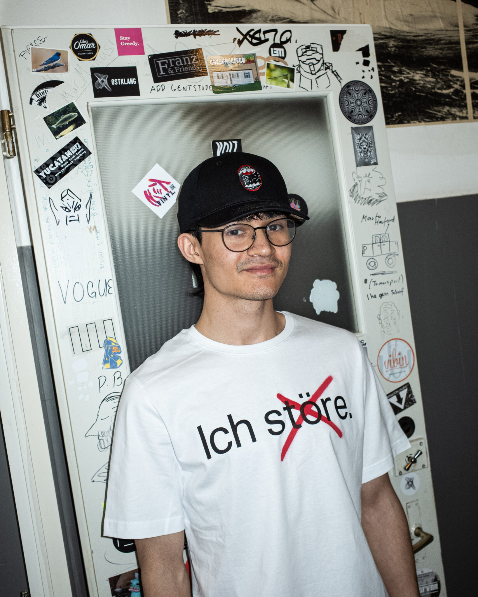 Ein junger Mann mit Brille, Mütze und einem weißen T-Shirt mit der Aufschrift "Ich störe" steht vor einer Tür, die mit Aufklebern und Zeichnungen bedeckt ist. Er lächelt leicht und schaut in die Kamera.