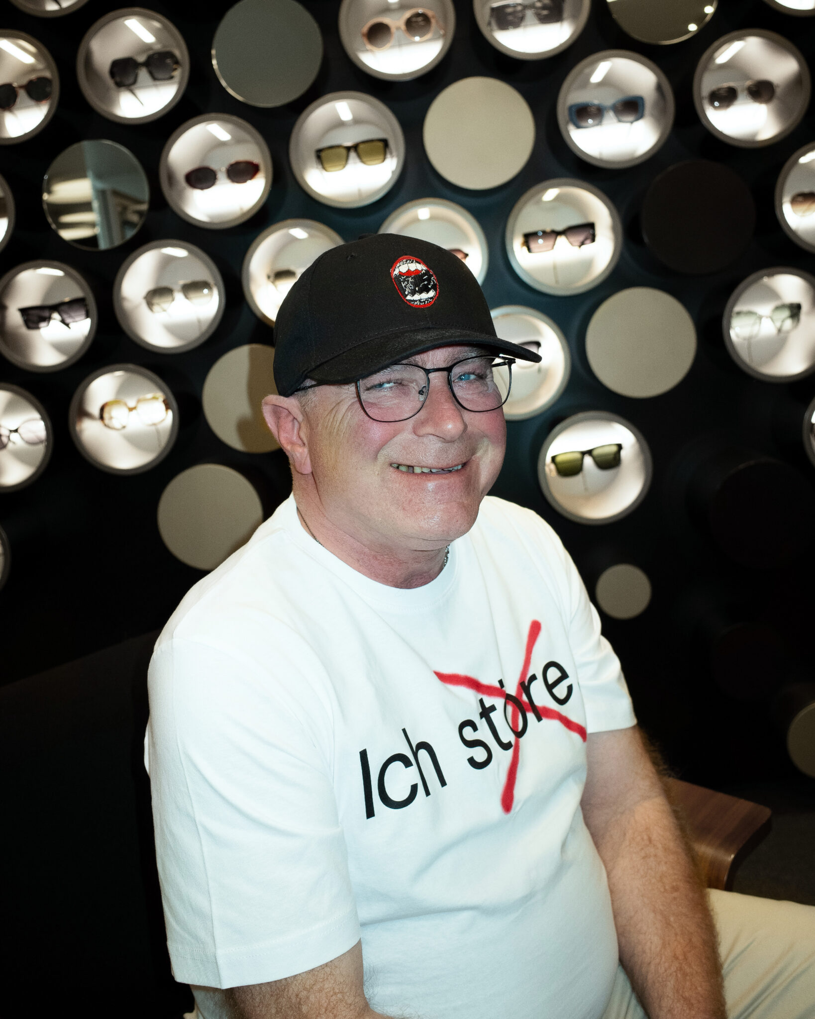 Ein lächelnder Mann mit Brille, schwarzer Mütze und weißem T-Shirt mit durchgestrichener Aufschrift "Ich store" sitzt vor einem Wanddisplay mit verschiedenen Sonnenbrillen in runden Rahmen.