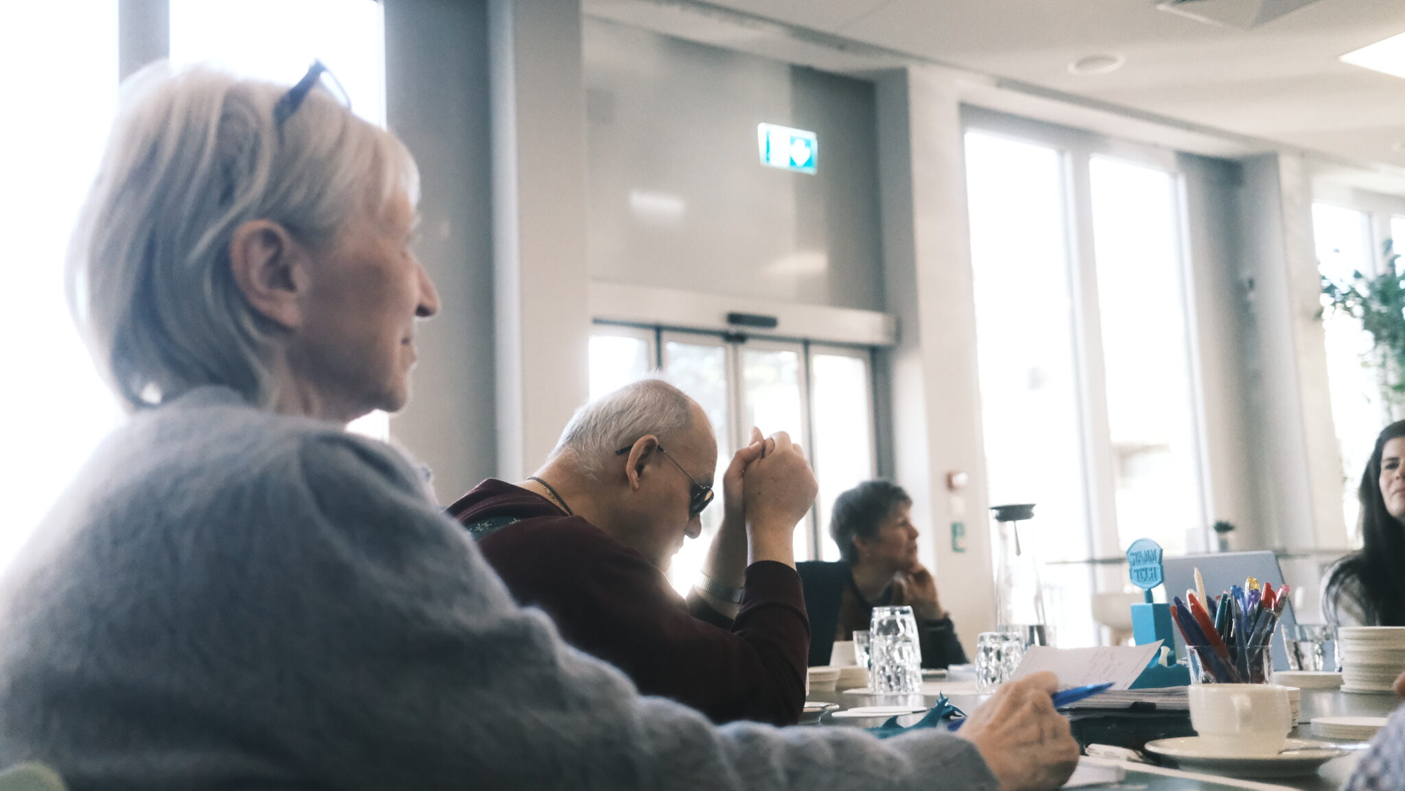Eine Gruppe von Menschen sitzt in einem hellen Raum um einen Tisch herum und unterhält sich. Eine Person scheint zu sprechen, während eine andere die Hände verschränkt und Tassen, Gläser und Papiere auf dem Tisch stehen hat.