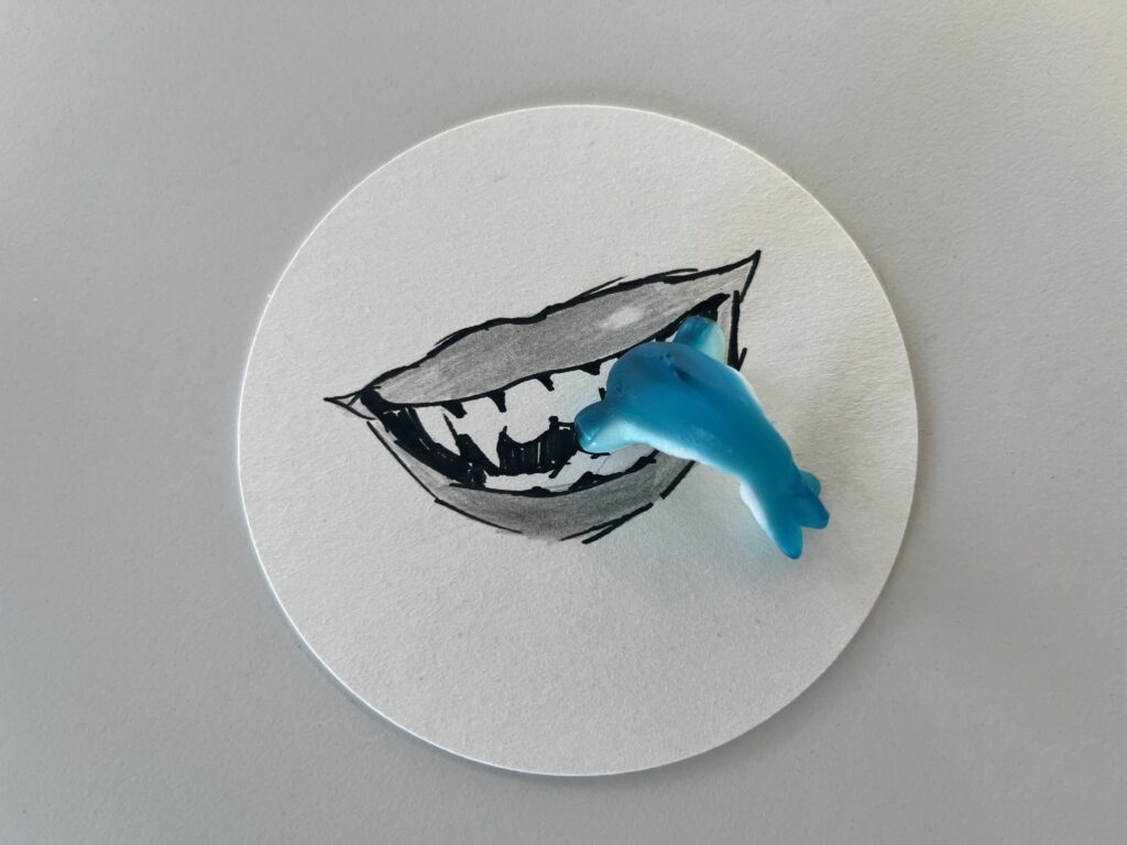 Ein rundes Stück Papier mit der Skizze eines offenen Mauls mit scharfen Zähnen. Ein kleiner blauer Spielzeug-Hai wird so platziert, dass er in das Maul zu schwimmen scheint.