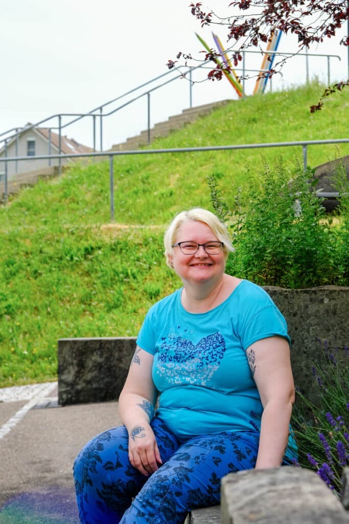 Eine lächelnde Person mit kurzen blonden Haaren, Brille und blau gemusterter Kleidung sitzt auf einer Steinbank im Freien, mit Grünpflanzen und Stufen im Hintergrund.