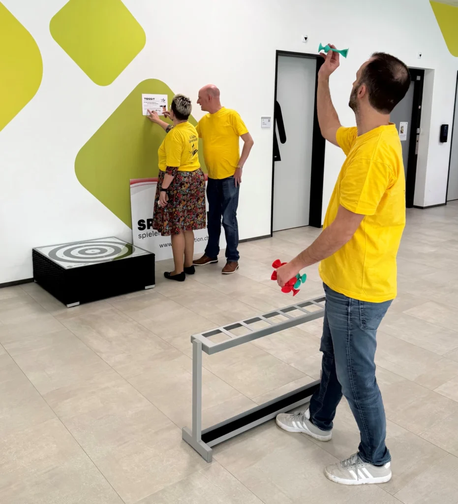 Drei Personen in gelben Hemden spielen ein Dartspiel in einer Halle. Ein Mann zielt auf die Dartpfeile, während eine Frau und ein weiterer Mann auf eine Zielscheibe an der Wand schauen. In dem Raum befinden sich eine schwarze Stufe und ein Metallgestell.