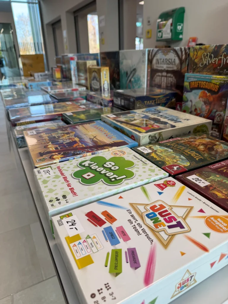 Auf einem langen Tisch stehen verschiedene bunte Brettspiele, darunter "Just One" und "So Kleever!" im Vordergrund, und im hinteren Teil des hellen Innenraums sind weitere Spielschachteln aufgereiht.