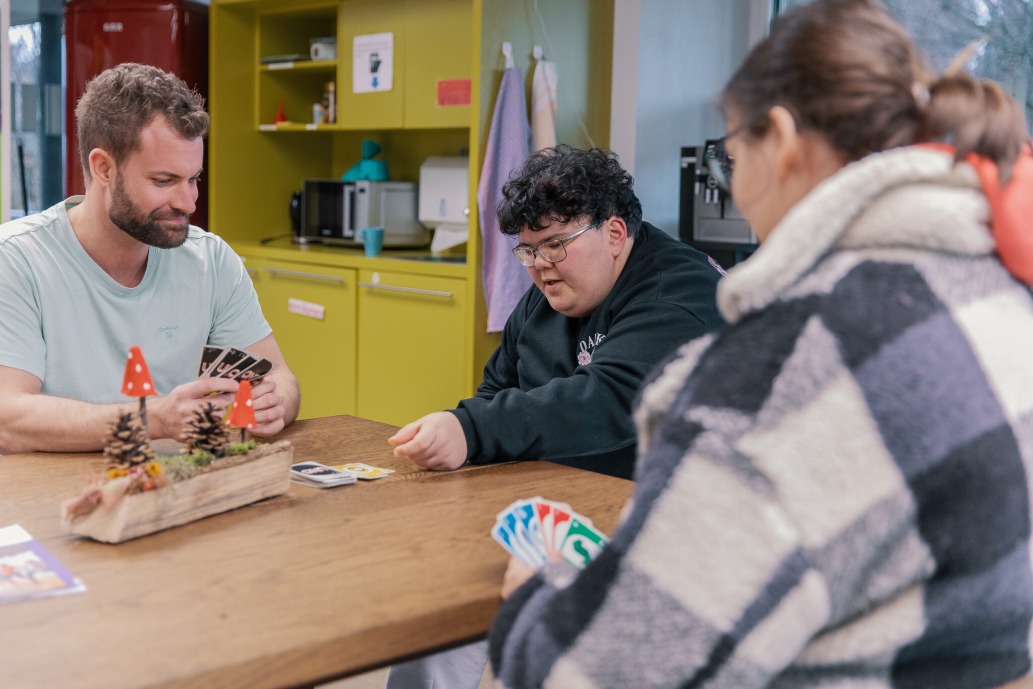 Drei Personen sitzen um einen Holztisch und spielen ein Kartenspiel in einem gemütlichen Raum mit gelben Schränken und Küchengeräten im Hintergrund. Eine Person legt eine Karte ab, während die anderen Karten halten.