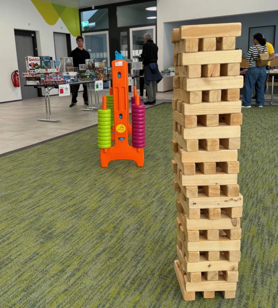 Ein hoher Turm aus hölzernen Jenga-Blöcken steht auf einem grünen Teppich, mit einem bunten Ringwurfspiel und Tischen mit Brettspielen im Hintergrund. Die Leute stöbern an den Tischen in einem hellen Innenraum.