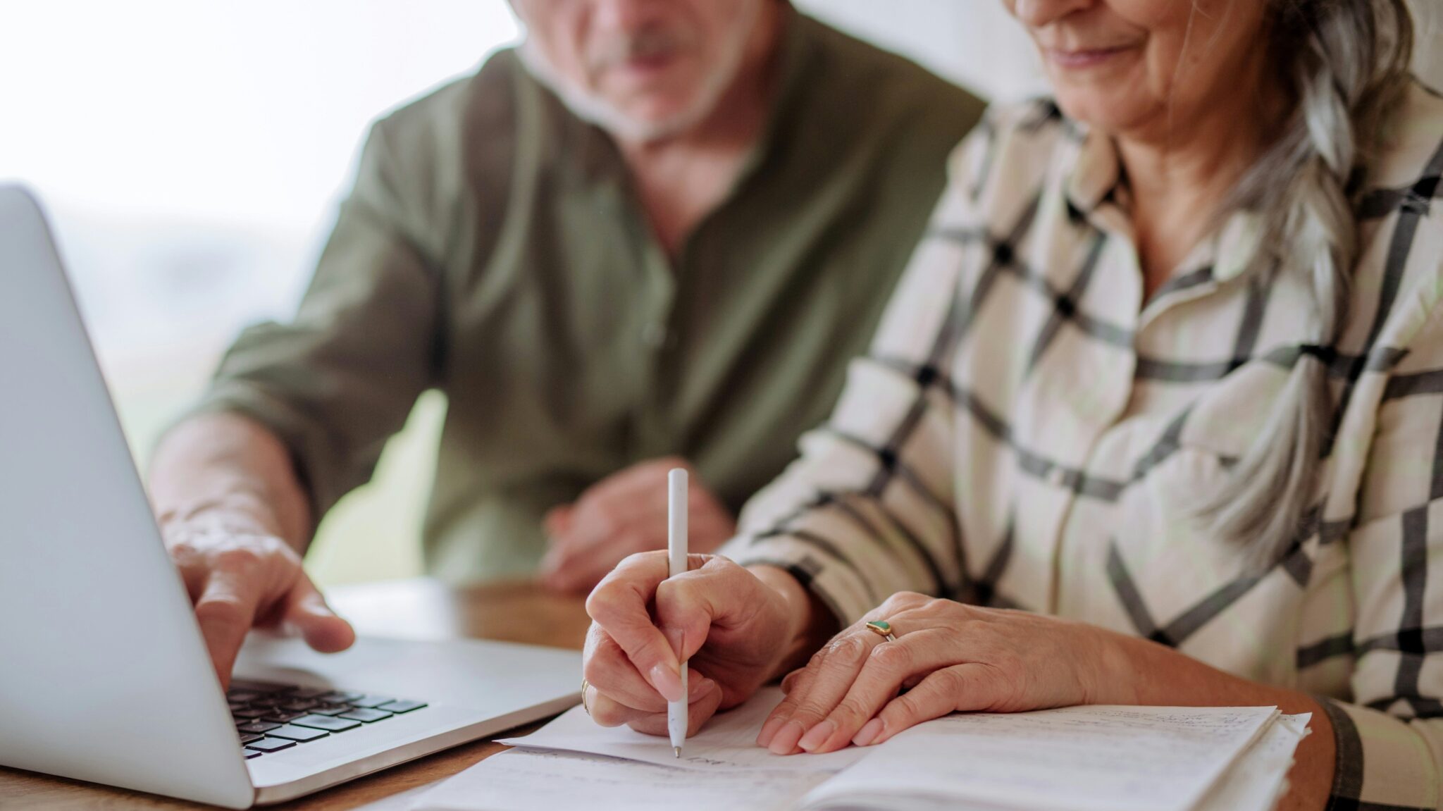 Zwei Personen sitzen an einem Tisch und benutzen einen Laptop. Eine Person schreibt mit einem Stift in ein Notizbuch, während die andere auf den Bildschirm des Laptops zeigt. Beide tragen legere Kleidung und arbeiten zusammen.