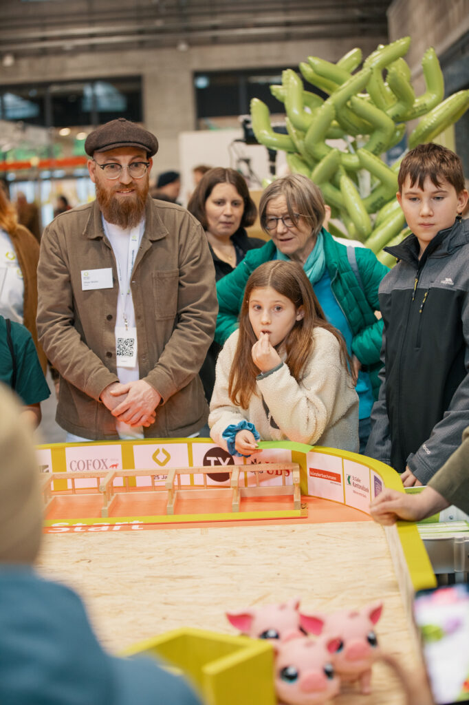 Eine Gruppe von Erwachsenen und Kindern beobachtet aufmerksam, wie sich kleine Spielzeugschweinroboter in einer gelb umzäunten Arena bei einer Indoor-Veranstaltung bewegen. Die Szene ist lebendig, mit farbenfrohen Dekorationen und konzentrierten Gesichtern.