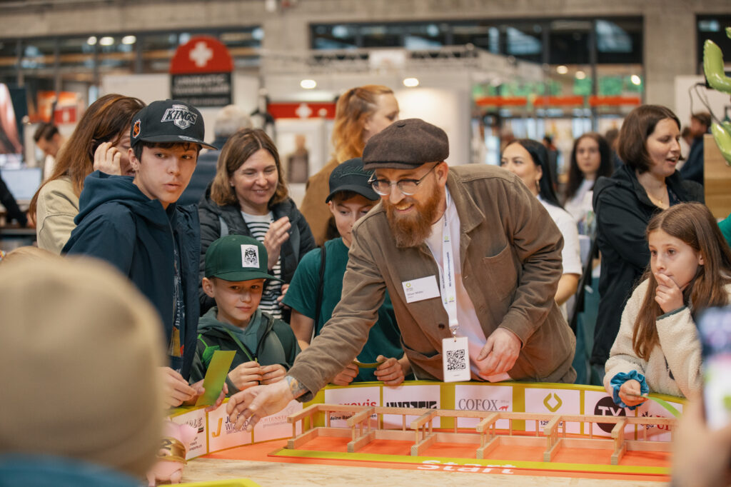 Ein bärtiger Mann mit brauner Mütze und Jacke führt Kinder bei einer belebten Indoor-Veranstaltung durch ein Tischspiel, während Zuschauer im Hintergrund lächelnd zuschauen.