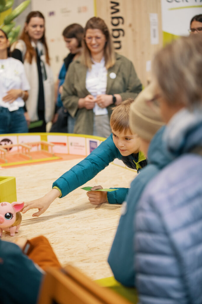Ein kleiner Junge in einer blauen Jacke greift nach einem schweinchenförmigen Spielzeug auf einem Tisch, während Erwachsene im Hintergrund bei einer Indoor-Veranstaltung zusehen und lächeln.