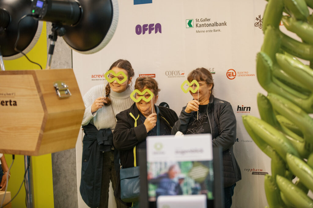 Drei Frauen posieren spielerisch mit grünen Papiermasken vor einer mit Logos bedeckten Fotokabinen-Kulisse bei einer Veranstaltung. Im Vordergrund sind eine große Fotografenleuchte und grüne Luftballondekoration zu sehen.