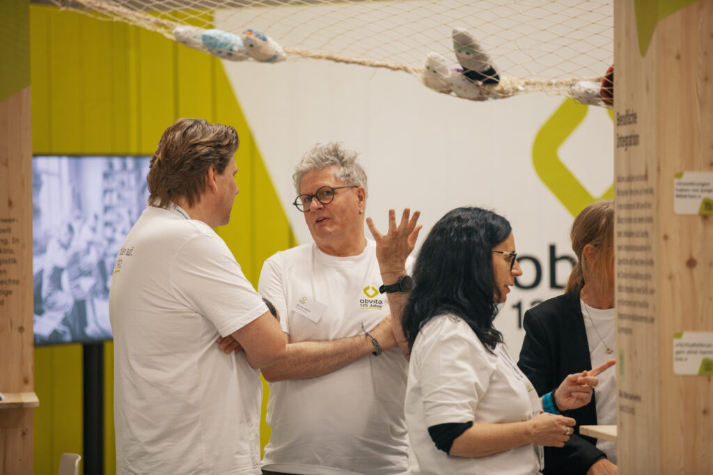 Vier Personen in weißen T-Shirts stehen und unterhalten sich an einem überdachten Veranstaltungsstand mit Holzkonstruktionen. Über ihnen hängt ein Netz mit ausgestopften Vögeln, und an den Wänden des Standes ist ein Informationstext angebracht.